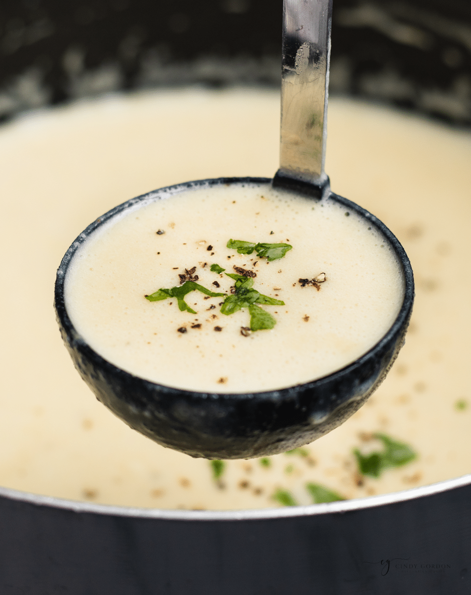 a black ladle holding up a serving of 4 ingredient creamy potato soup, garnished with black pepper and herbs.