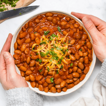 hands holding a large bowl of ranch style beans with cheddar and cilantro on top.