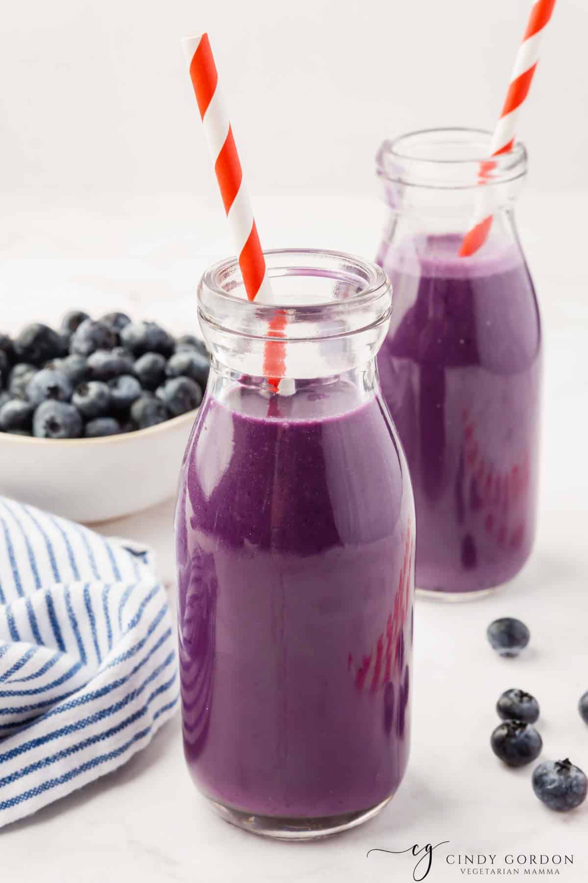 Two small glass jars filled with purple blueberry milk. There are red and white straws in each jar, and a bowl of blueberries in the background. 