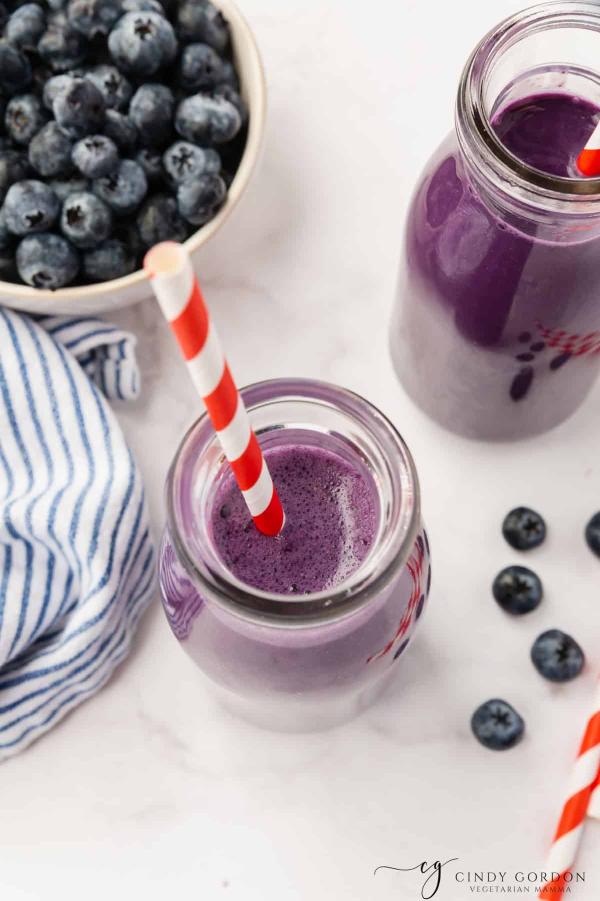 vertical photo showing 2 small bottles of blueberry milk with straws in them from overhead, surrounded by a bowl of blueberries and some loose blueberries all on a white surface