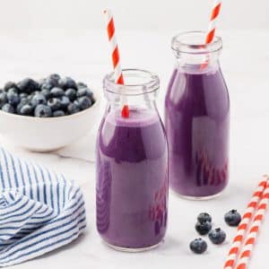 Two small glass jars filled with purple drink. There are red and white straws in each jar, and a bowl of blueberries in the background.