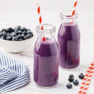 Two small glass jars filled with purple drink. There are red and white straws in each jar, and a bowl of blueberries in the background.