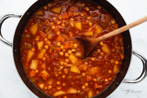 a soup pot with broth, tomatoes, veggies, beans, a wooden spoon is stirring.