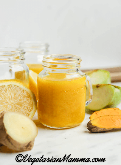 A small mason jar mug filled with orange ginger turmeric juice. Around the jar are pieces of apples, lemon, ginger, and turmeric root