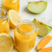 A small mason jar mug filled with orange ginger turmeric juice. Around the jar are pieces of apples, lemon, ginger, and turmeric root