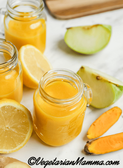A small mason jar mug filled with orange ginger turmeric juice. Around the jar are pieces of apples, lemon, ginger, and turmeric root