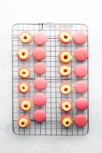 A mesh cooling rack holding dark pink macaron shells with lemon and strawberry filling piped on to half.