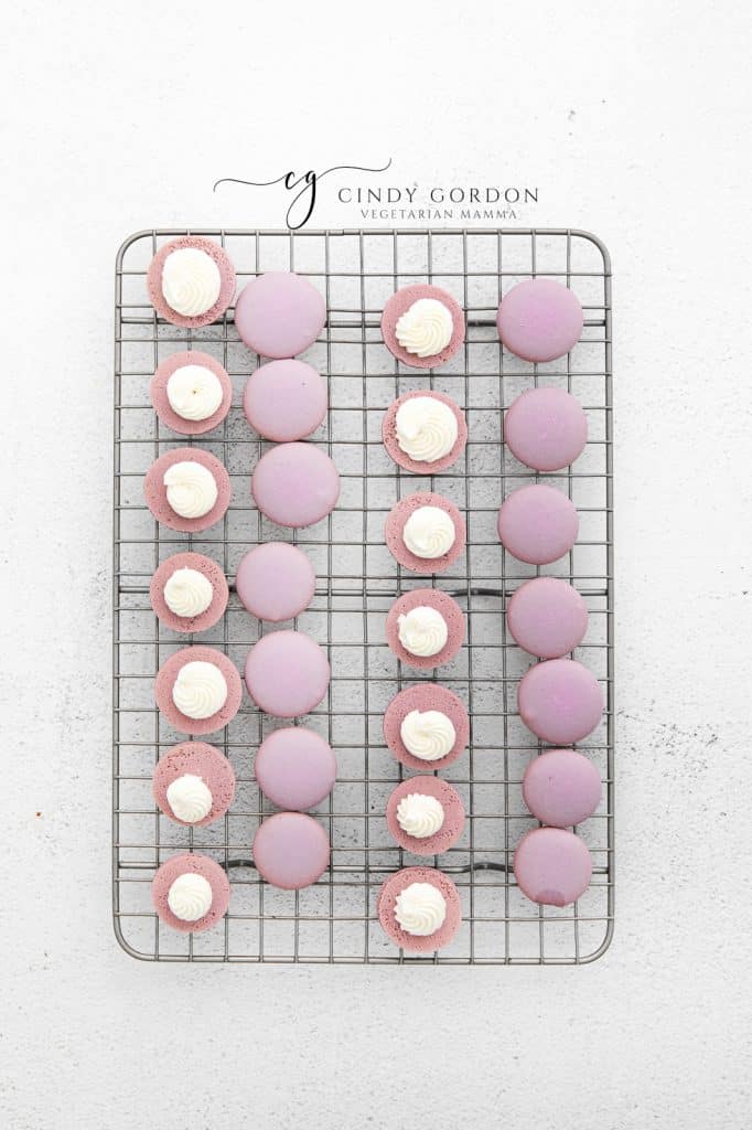 lavender macaron shells on a wire rack with frosting added to half of them.