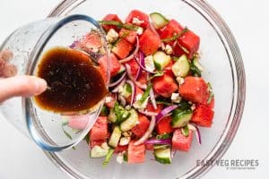 balsamic dressing being poured over a bowl of watermelon and vegetables.
