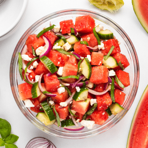 a bowl of watermelon basil salad, viewed from above. Around the bowl are wedges of melon and basil leaves.