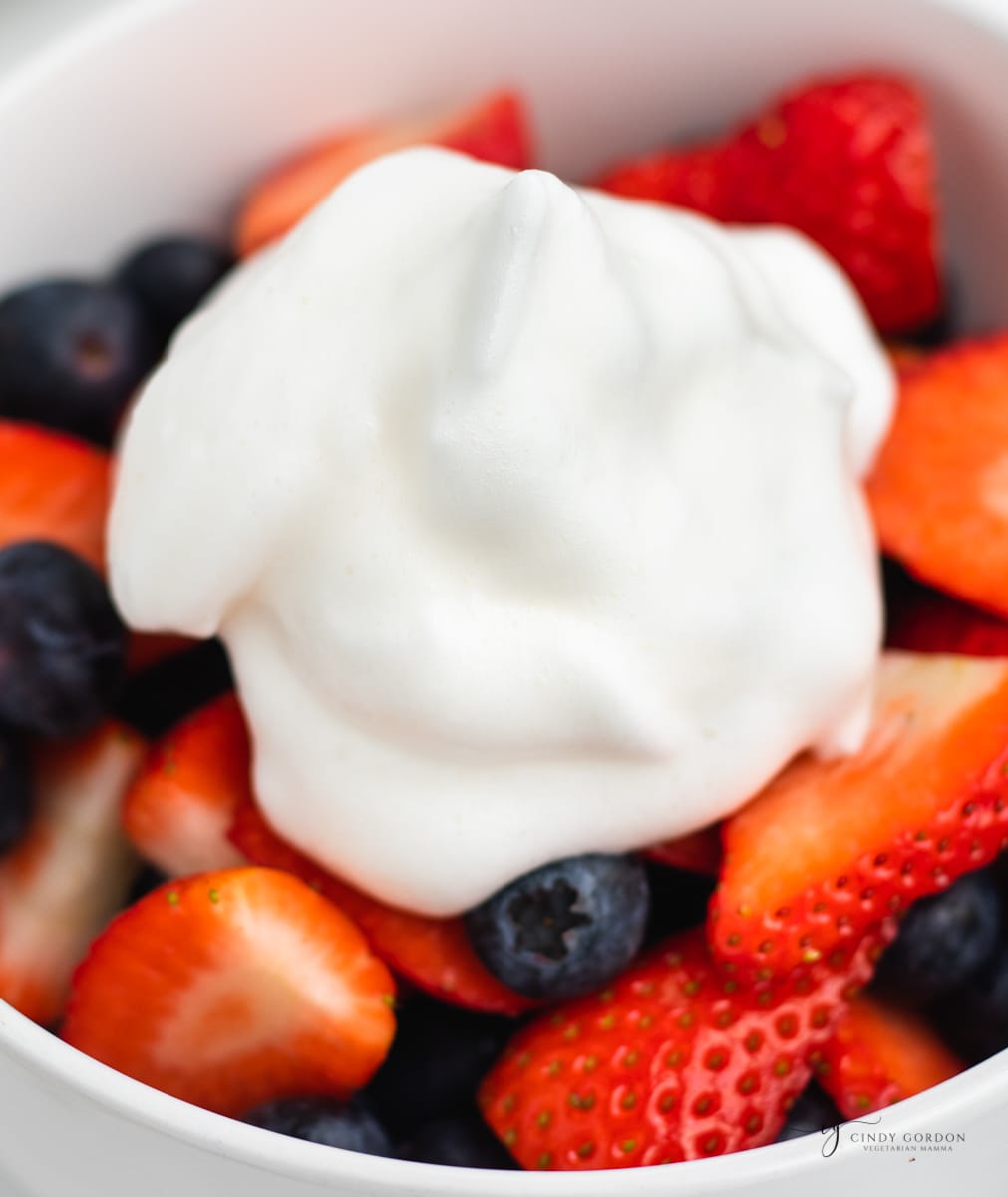 a dollop of aquafaba whipped cream on top of a bowl of strawberries and blueberries. 