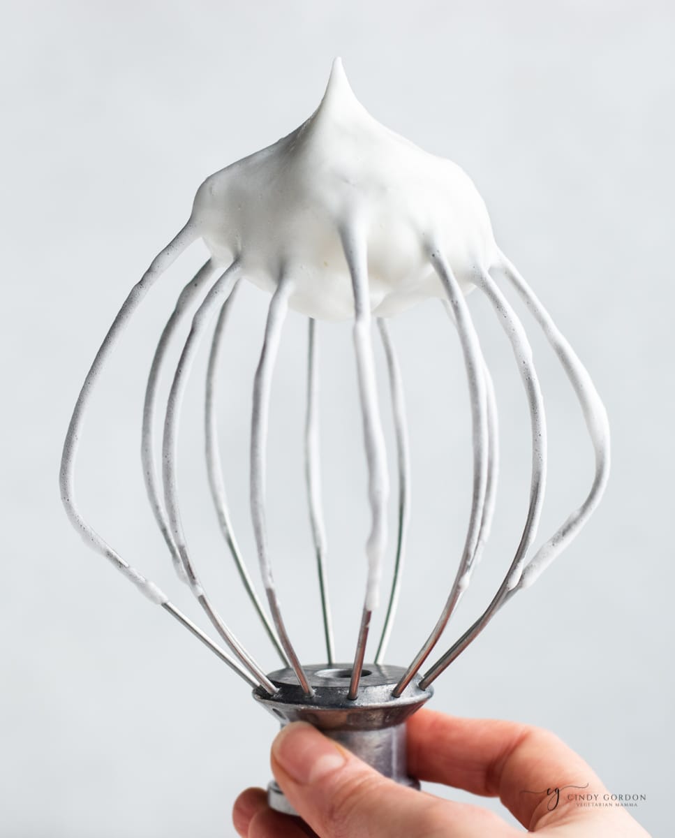a mixer whisk held up to show aquafaba whipped into stiff peaks.