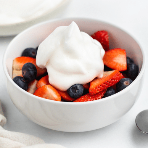 a white bowl of sliced strawberries and blueberries topped with a dollop of aquafaba whipped cream.
