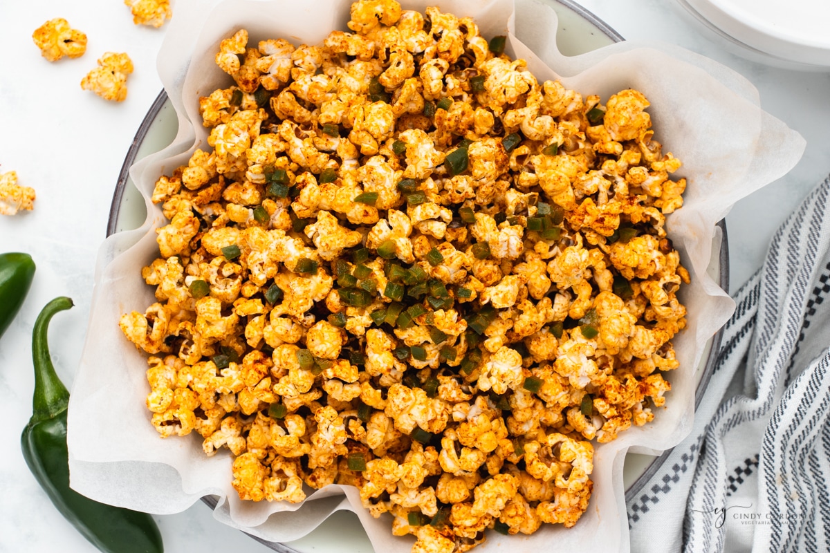 a bowl lined with a towel and filled with spicy popcorn, topped with diced jalapeno