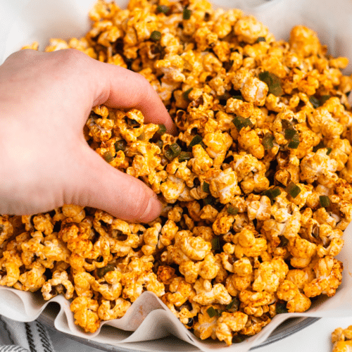 a bowl of popcorn coated in chili powder and topped with diced jalapeno. A hand is grabbing popcorn from the bowl.