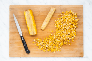 A wooden cutting board holding a sharp knife and two ears of corn that are in the process of sliced to remove the kernels.