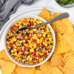 a white bowl of roasted corn pico on a plate of corn chips.