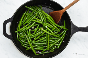 a cast iron skillet of green beans with garlic being stirred with a wooden spoon.