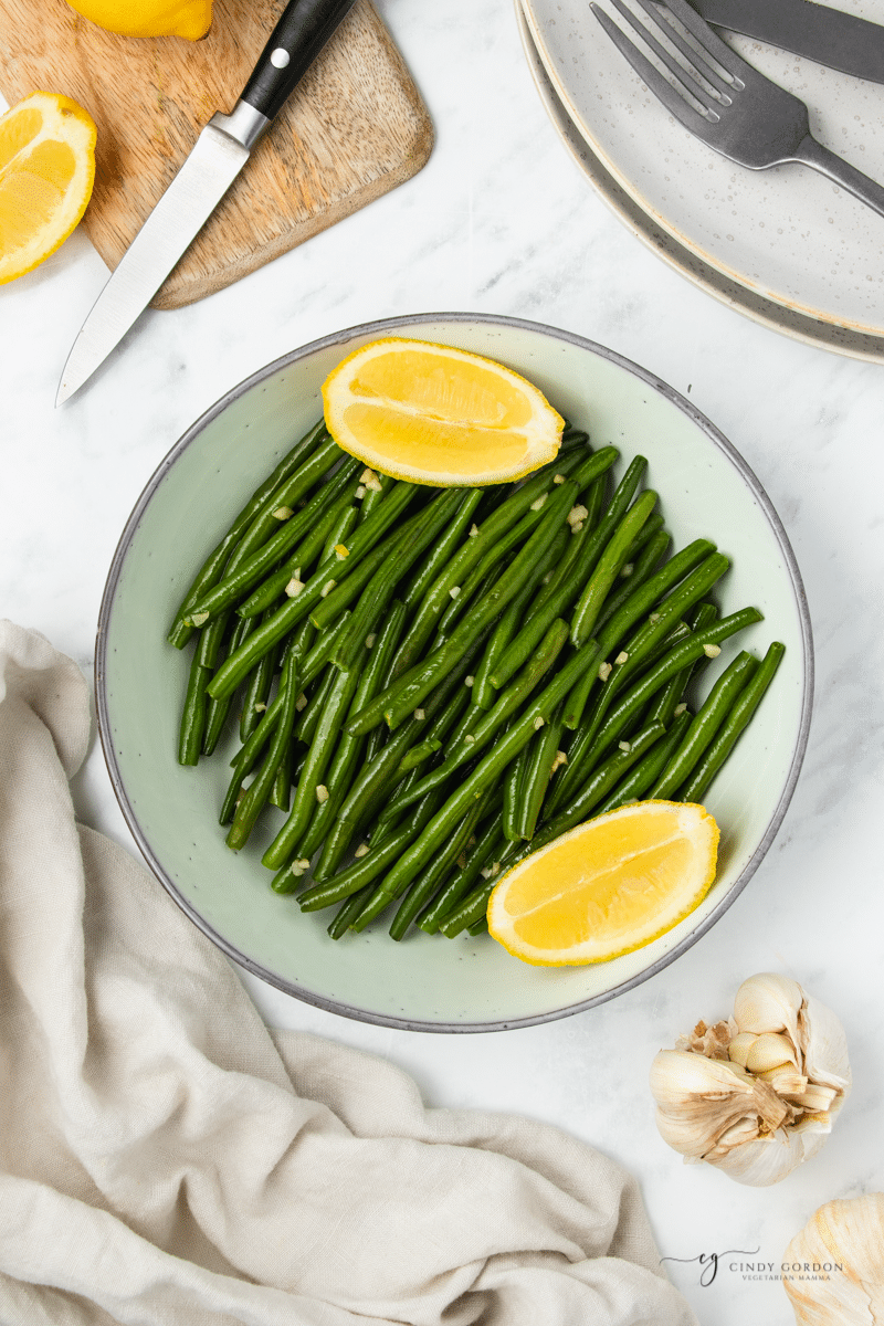 a serving bowl of sauteed green beans with garlic and two lemon wedges. 