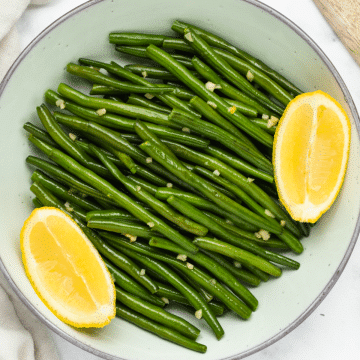 a white serving bowl filled with sauteed green beans with garlic and lemon wedges.
