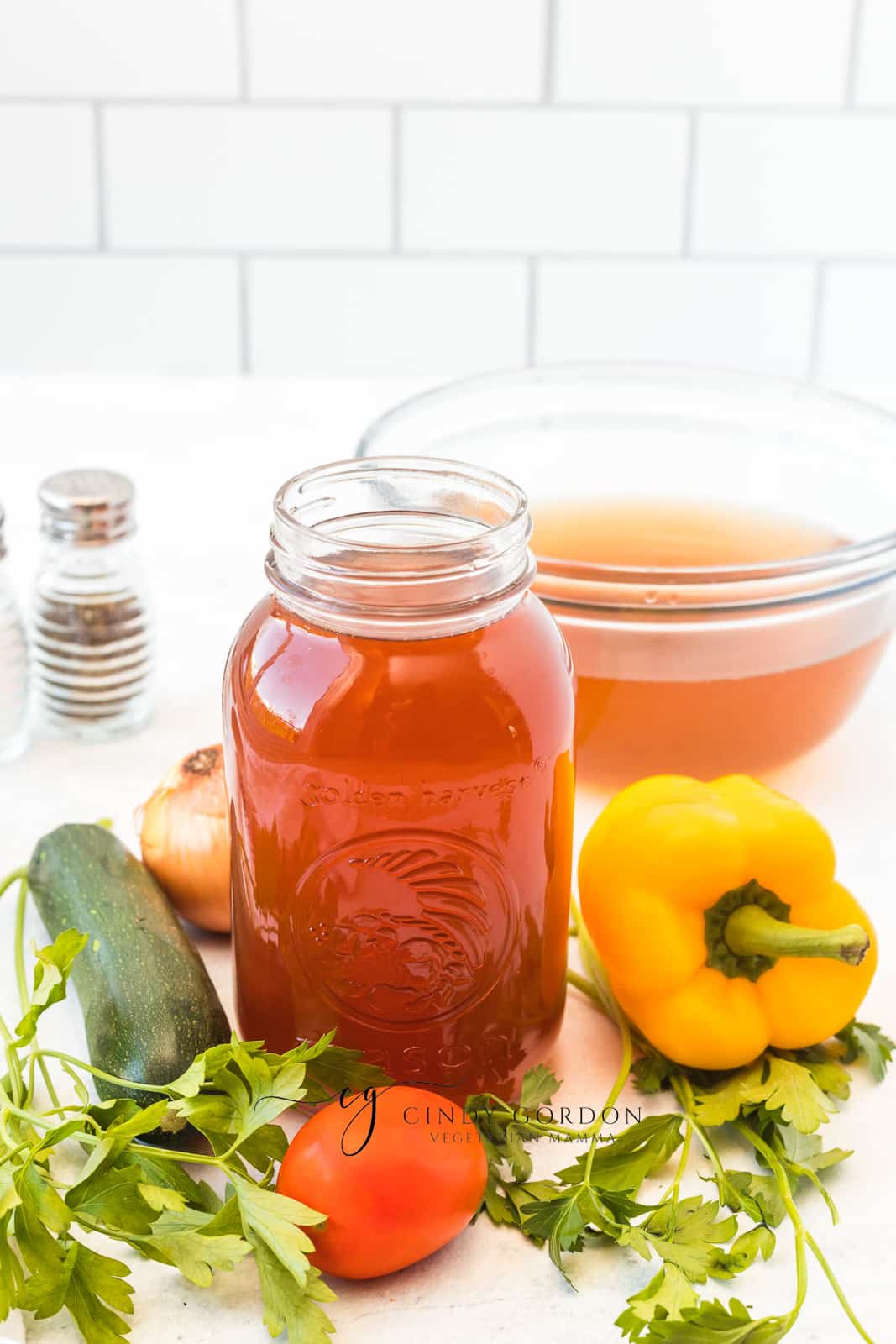 red broth in a mason jar next to zucchini,k herbs, tomato, and a yellow pepper.