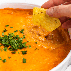 a white dish of vegan buffalo chicken dip with chopped herbs on top. A hand is dipping a chip into the dip.