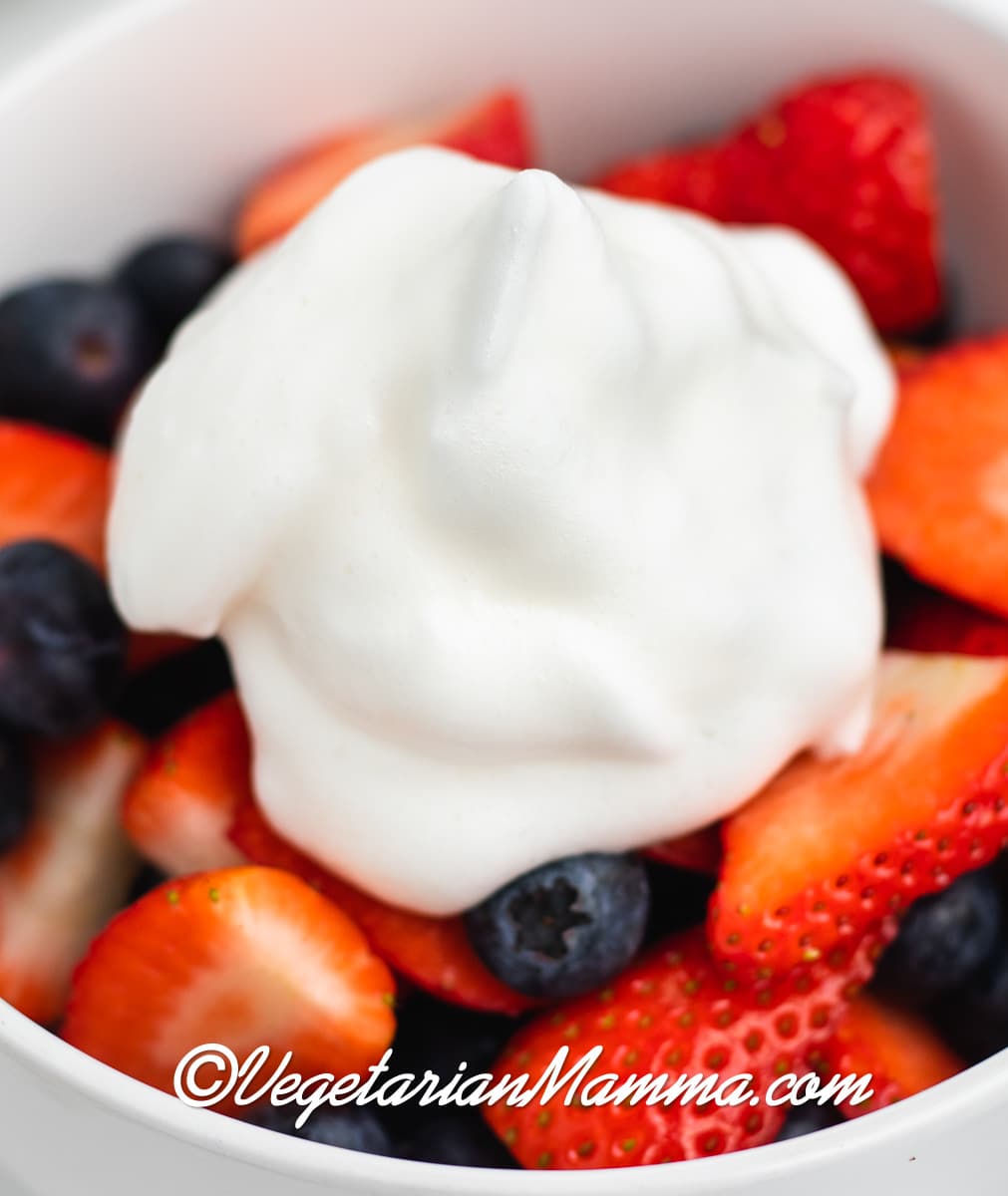 a white bowl of sliced strawberries and blueberries topped with a dollop of aquafaba whipped cream.