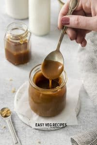 a hand holding a spoon that has dipped into a small jar of vegan caramel sauce.
