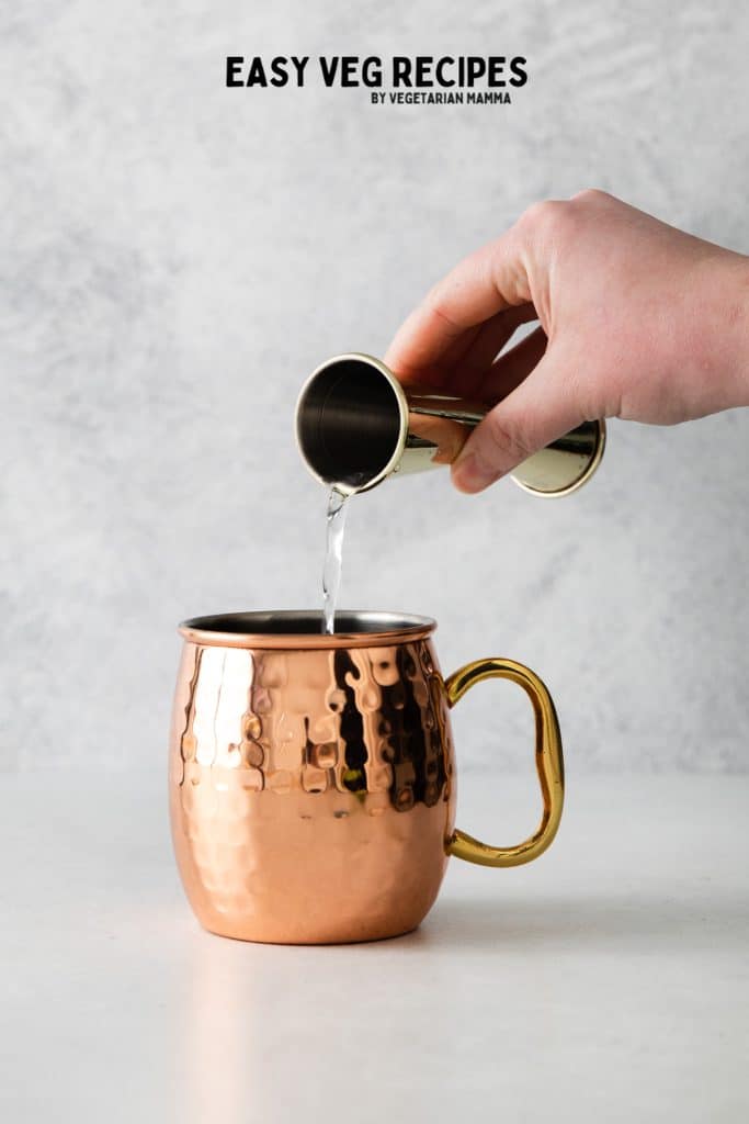 a hand holding a jigger, pouring gin into a chilled copper mug.