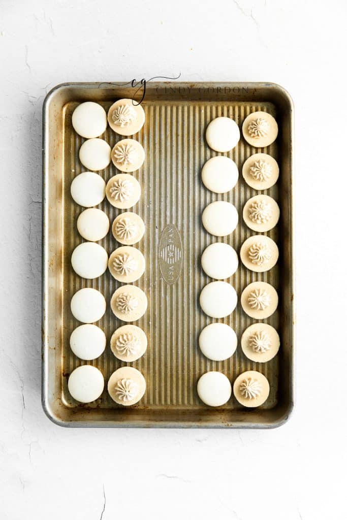 baked macaron shells on a tray. Half are topped with filling.