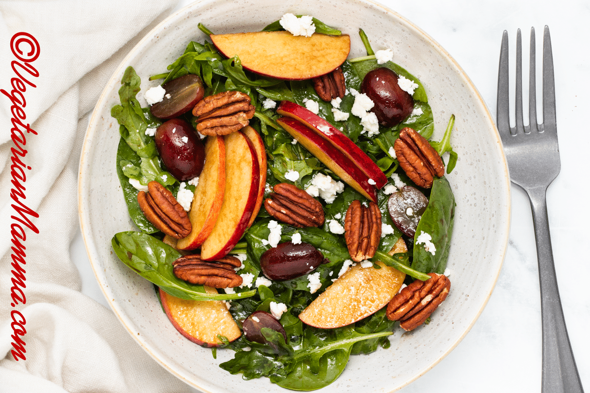 A green salad with apples, grapes, pecans, and feta cheese, on a plate, next to a fork. 