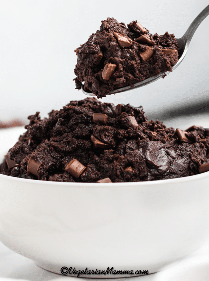 a white bowl filled with dark chocolate edible brownie batter. A spoon is lifting out a spoon full of it.